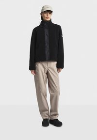 Giacca in pile nera con collo alto e pannello frontale; abbinata a pantaloni beige e calzature nere; il modello indossa un berretto chiaro.