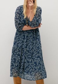 Žena v námořnickém modrém květinovém midi šatech s průhlednými rukávy, hlubokým výstřihem do V a světle hnědými kozačkami po kolena, stojí s jednou rukou blízko brady.