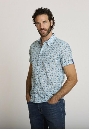 Homme aux cheveux bouclés et à la barbe portant une chemise à manches courtes à boutons avec un motif floral bleu et un jean bleu, les mains dans les poches.