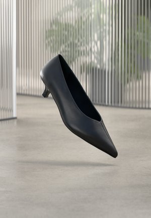 Chaussure noire à talon kitten à bout pointu flottant au-dessus d'un sol en béton lisse avec des panneaux translucides verticaux en arrière-plan.