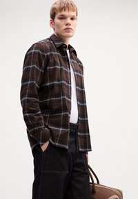 Junger Mann in einem braun karierten Hemd über einem weißen T-Shirt, dunklen Hosen, mit einer Hand in der Tasche, hält eine braune und beige Tasche.