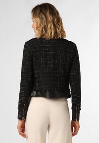 Schwarze Tweedjacke mit strukturiertem Finish, Lederakzenten am Saum und an den Bündchen sowie Reißverschlussbündchen, von hinten betrachtet.