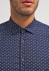 Camicia blu con motivo a fiori bianchi piccoli, colletto standard, chiusura frontale con bottoni e tessuto testurizzato. Presenta bottoni marroni.