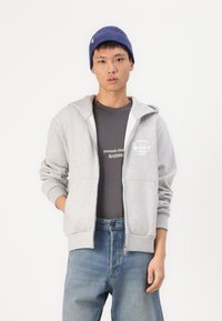 Sweat à capuche gris avec une poche avant et des poignets côtes, porté sur un t-shirt gris foncé. Le modèle l'associe avec un jeans en denim bleu et un bonnet bleu marine.