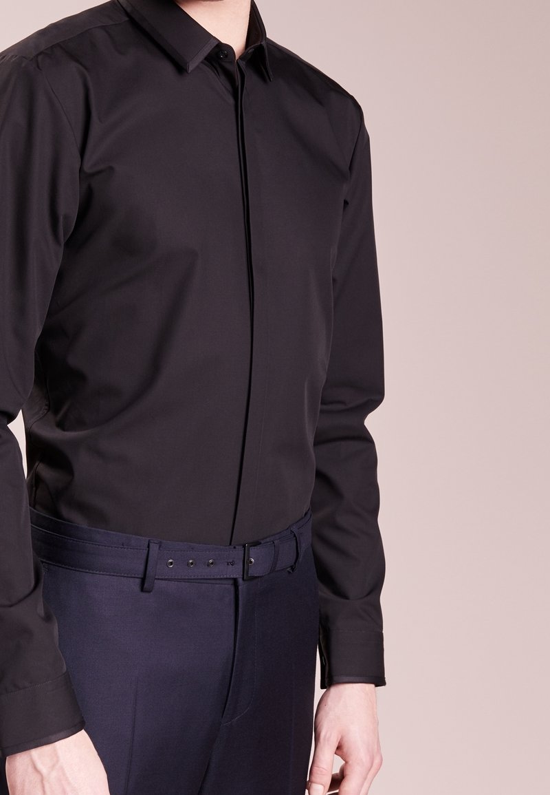 Homme portant une chemise de ville noire ajustée rentrée dans un pantalon de costume bleu marine avec une ceinture assortie, visible des épaules aux hanches.