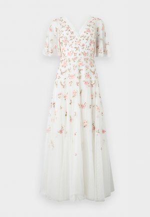 Femme portant une robe blanche longueur cheville avec des manches bouffantes transparentes et une broderie florale rose, assortie à des talons blancs pointus.