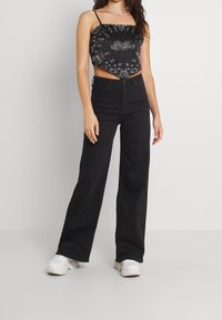 Svart kort topp med vita mönster, kombinerad med högmidjade svarta vida jeans och vita sneakers. Modellen står med händerna på höfterna.