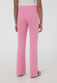 Pantaloni a zampa d'elefante rosa con una texture liscia, caratterizzati da fessure laterali all'orlo e una cintura elastica, indossati con calzature di colore blu chiaro.