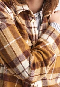 Camisa de franela a cuadros con colores amarillo, marrón y crema. Textura suave con cuello curvo y botones dorados. Tela en capas visible.