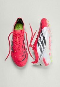 Ζευγάρι κόκκινων και λευκών παπουτσιών ποδοσφαίρου με μαύρες λεπτομέρειες και κορδόνια ξεκούμπωτα, τοποθετημένα δίπλα-δίπλα σε ανοιχτό γκρι φόντο.