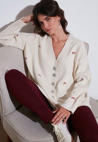Cardigan color crema con ricamo a forma di fiocco rosso, cinque grandi bottoni argento, design a V, abbinato a pantaloni bordeaux e sneakers bianche.