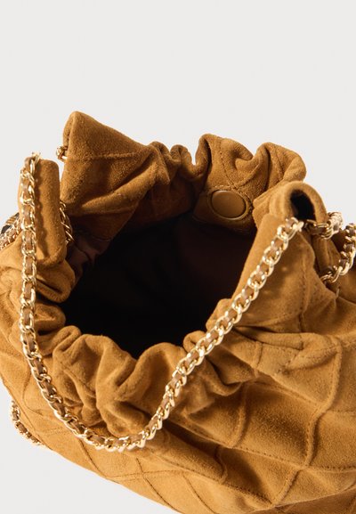Ruskea mokkasuede-narupussukka, jossa on poimutettu muotoilu, kultaketjuolkapää ja pehmeä, teksturoitu sisus. Varustettu napilla suljettavalla kiinnityksellä.
