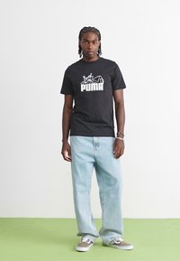Sort bomulds T-shirt med hvid "PUMA" logo, lyseblå baggy jeans og grå sko med hvide accenter, stående på et grønt gulv.