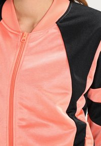 Růžová a černá bunda na zip s hladkým materiálem, kontrastním designem panelů a žebrovaným límcem. Je vidět detail předního zipu.