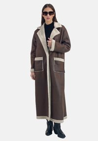 Lange bruine jas met een pluizige witte kraag en bijpassende zoom, met twee voorzakken en een losse pasvorm. Draagt over een zwarte top.