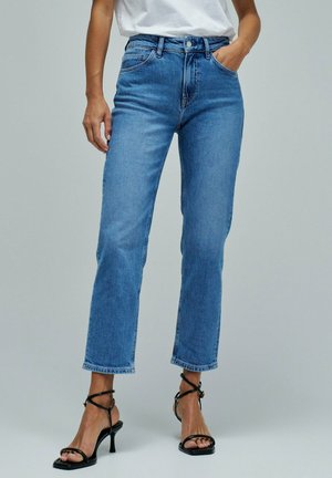 Mujer con vaqueros azules de talle alto y pierna recta y sandalias negras de tacón con tiras, de pie frente a un fondo liso.