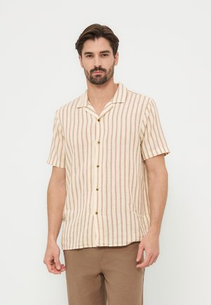 Άνδρας που φοράει μπεζ και καφέ πουκαμίσα με κάθετες ρίγες, κοντομάνικη με κουμπιά, και καφέ παντελόνι, στέκεται μπροστά σε απλό λευκό φόντο.