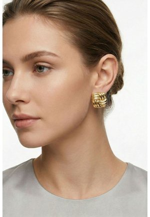 Ženska s svetlo rjavimi lasmi, zbrkanimi nazaj, nosi veliko zlato teksturirano kvadratno obrobo in svetlo sivo majico na enobarvni podlagi.