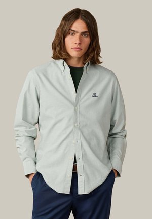 Hombre con cabello castaño hasta los hombros, con camisa gris claro abotonada con logo y pantalones azul oscuro, manos en los bolsillos, fondo neutro.