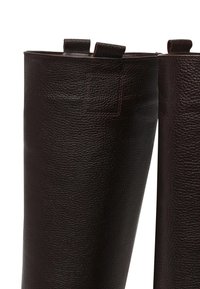 Bruine leren laarzen tot aan de knie met een gestructureerd oppervlak. Voorzien van twee lusjes aan de bovenkant voor gemakkelijk dragen. Close-up, zonder zolen.