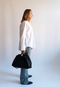 Mujer con camisa blanca y vaqueros sosteniendo una gran bolsa negra, de pie de lado contra un fondo liso y claro.