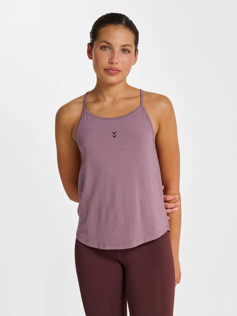 Jeune femme aux cheveux tirés en arrière portant un débardeur mauve et des leggings violet foncé, debout devant un fond blanc uni.