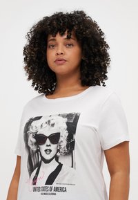 Hvit T-skjorte med en stor svart-hvitt grafikk av en kvinne med solbriller. Teksten under sier "MARILYN UNITED STATES OF AMERICA."