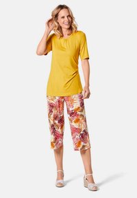 Haut jaune à manches courtes associé à un pantalon capri blanc à motifs tropicaux colorés. La mannequin porte des sandales à talons blanches.