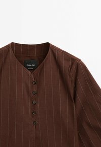 Brun skjorte med opretstående krave og hvide striber. Har en v-hals og seks sorte knapper foran. Lavet af struktureret stof.