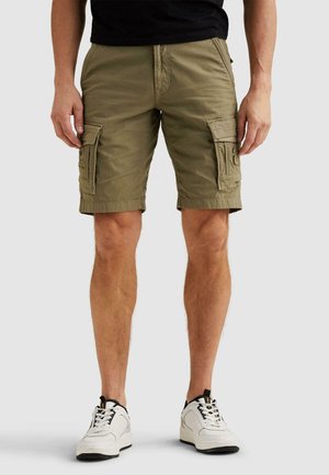 Mand iført olivengrønne cargobukser og hvide sneakers, stående mod en ensfarvet lys baggrund.