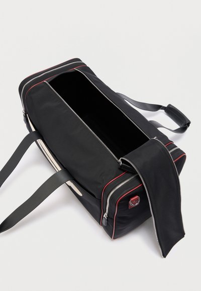 Μαύρη ορθογώνια τσάντα duffel με κόκκινη διακοσμητική ραφή, με μερικώς ανοιγμένο κύριο διαμέρισμα με φερμουάρ, διπλές μαύρες λαβές και ρυθμιζόμενο ιμάντα ώμου σε λευκό φόντο.