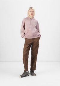 Lys rosa hettegenser med frontlomme, kombinert med brune bukser og grå sko. Hettegenseren har snøring og et merke som detalj.
