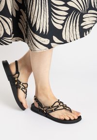 Black sandals with a flat sole and intricate braided straps featuring gold accents and an adjustable buckle for a secure fit.
