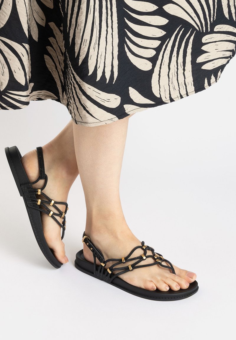 Black sandals with a flat sole and intricate braided straps featuring gold accents and an adjustable buckle for a secure fit.