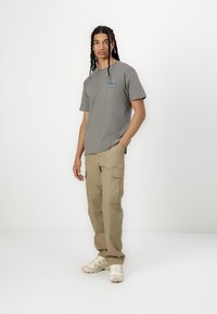 Graues Baumwoll-T-Shirt mit Grafiken auf der Vorderseite, kombiniert mit beigen Cargo-Hosen, die über mehrere Taschen und eine lockere Passform verfügen. Die Schuhe sind hellfarbige Sneaker.