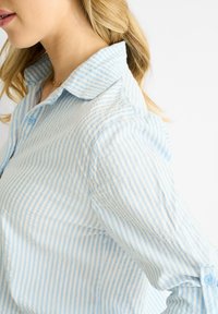 Camicia button-up a righe blu chiaro e bianco con colletto, realizzata in tessuto morbido e con maniche arrotolate. Texture sottile con un design classico.