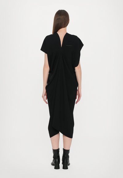 Black dress with a draped design, short sleeves, and a V-shaped back. Material appears soft and textured. Worn with black ankle boots.