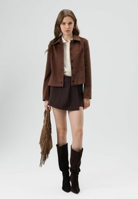 Bruine wollen jas met zwarte knopen, witte gebreide blouse, donkerbruine mini-rok en kniehoge bruine suède laarzen met franjes tas.