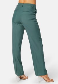 Hoge taille, wijde broekspijpen in teal stof, voorzien van achterzakken en een gladde textuur, gecombineerd met doorzichtige haksandalen.