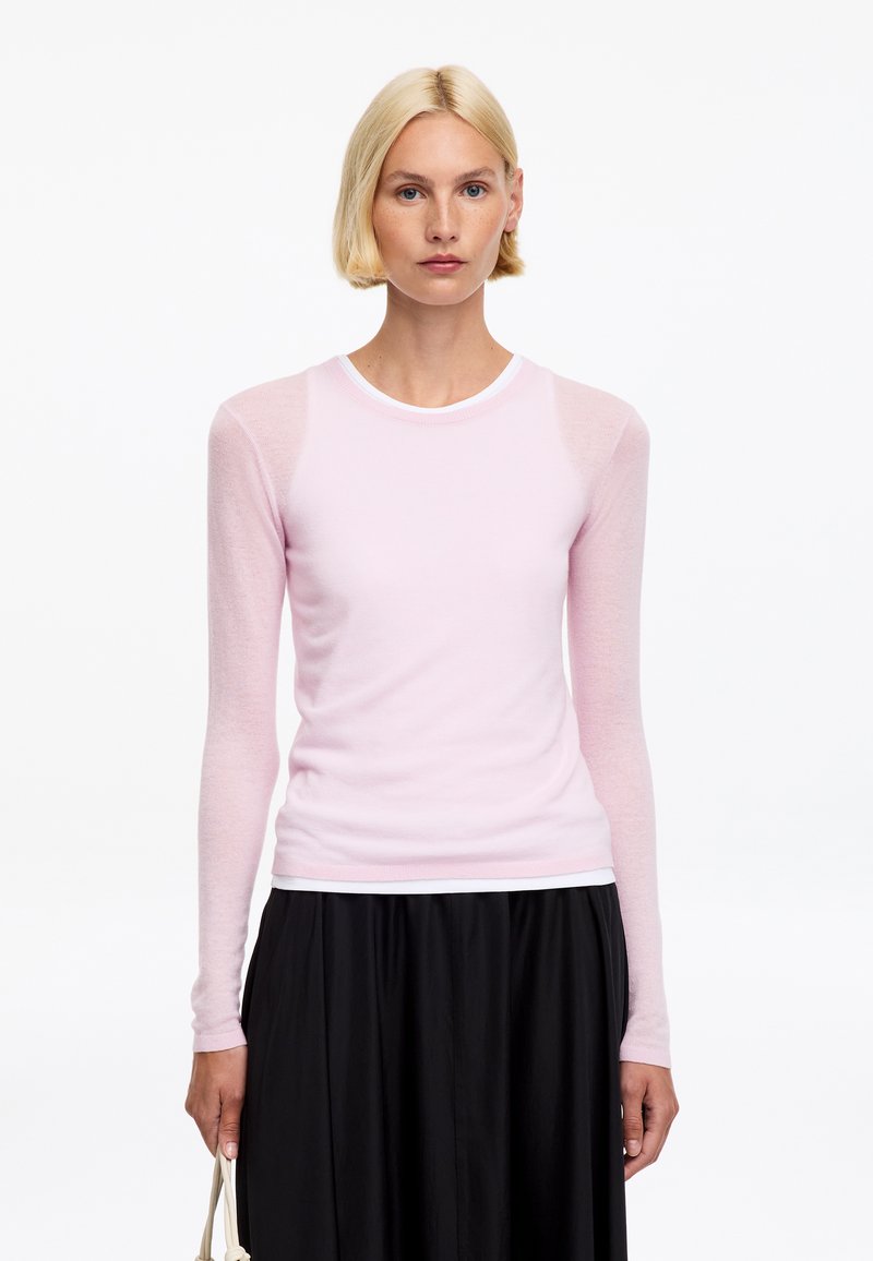 Femme blonde portant un haut à manches longues rose clair et une jupe noire, tenant un sac à main beige sur un fond blanc.