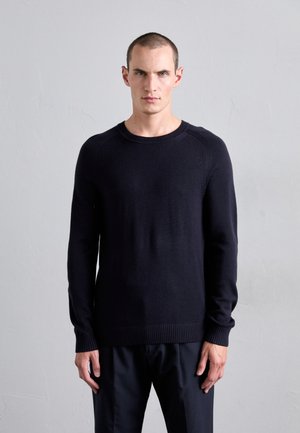 Mann mit kurzen Haaren, der einen schlichten schwarzen Langarmpullover und schwarze Hose trägt und gegen eine hellgraue strukturierte Wand steht.