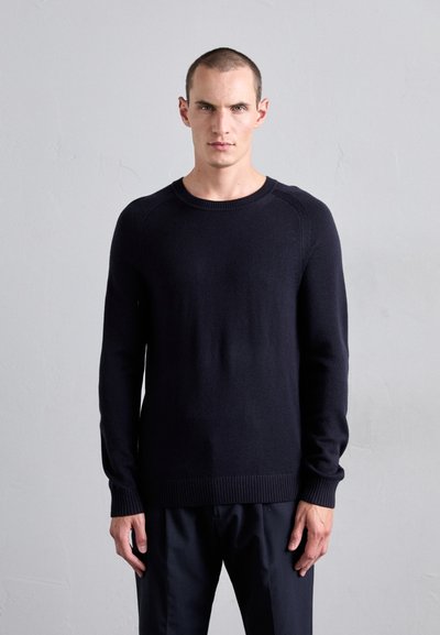 Man with short hair wearing a plain black long-sleeve sweater and black pants, standing against a light gray textured wall.