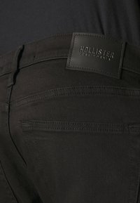 Juodi džinsai su lygiu paviršiumi, su siūtu juosmeniu ir stačiakampiu odiniu ženkliuku su įspaustu užrašu "HOLLISTER CALIFORNIA."