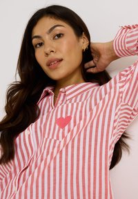 Camisa de botones a rayas en rosa y blanco con un parche de corazón rojo en el bolsillo del pecho. La tela parece suave y tiene un corte holgado.