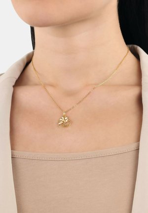 Collana d'oro con un ciondolo a forma di cuore decorato con un design di albero, su una delicata catena. Indossata sopra un top beige con un collo semplice.