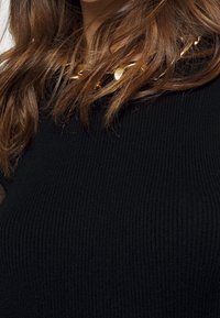 Svart ribbad tröja med rundad hals och ett guldhalsband med rundade, reflekterande detaljer. Strukturell detalj på tyget.