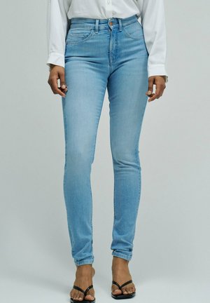 Mujer con vaqueros ajustados azul claro, camisa blanca abotonada y sandalias negras abiertas en los dedos, de pie frente a un fondo liso.