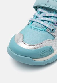 Zapatilla turquesa con una puntera metálica plateada, acentos azules texturizados, cordones con estampado y detalles florales en el lateral.