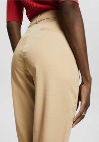 Pantaloni beige a vita alta con texture liscia, caratterizzati da una singola tasca posteriore e una silhouette aderente che si estende fino alla caviglia.