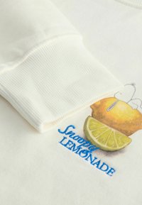 Sweatshirt couleur crème avec des poignets côtelés, affichant un motif de citron et de lime accompagné du texte « Snoopy Lemonade ». 